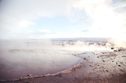 A geothermal landscape with a pool of water in the foreground surrounded by rock and soil. Steam rises gently from the surface, blending with the cloudy sky. The horizon shows distant mountains under a pale blue sky, creating a serene and expansive view.