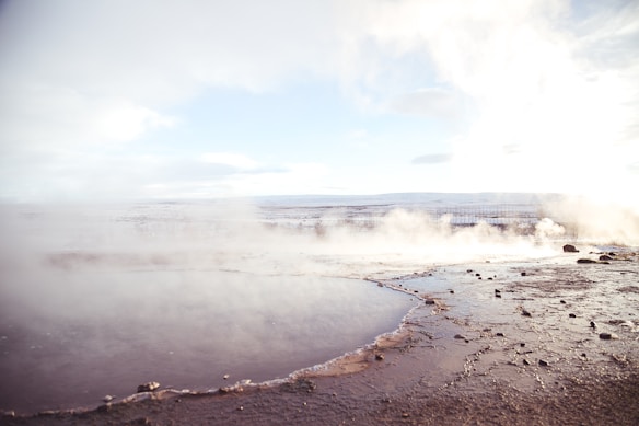 A geothermal landscape with a pool of water in the foreground surrounded by rock and soil. Steam rises gently from the surface, blending with the cloudy sky. The horizon shows distant mountains under a pale blue sky, creating a serene and expansive view.