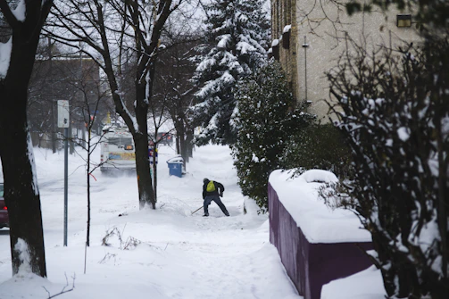 Close-up of a shovel pushing thick snow off a sidewalk beside a cozy home.