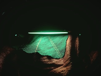 A bright, fluorescent green line of light illuminates textured fabric with intricate stitching. The fabric appears as a rugged green surface with prominent stitches, contrasted by deep shadows.