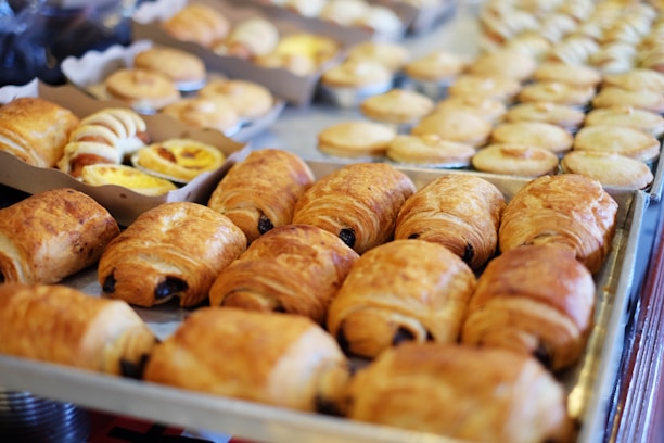 A variety of freshly baked pastries are displayed on metal trays. The selection includes croissants and other baked goods with golden brown crusts, arranged neatly in rows. The background features more pastries of various shapes and sizes.