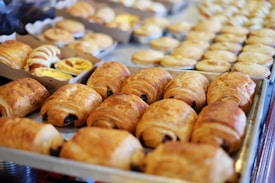 A variety of freshly baked pastries are displayed on metal trays. The selection includes croissants and other baked goods with golden brown crusts, arranged neatly in rows. The background features more pastries of various shapes and sizes.