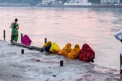 A group of people, mostly women dressed in brightly colored traditional garments, are sitting on a riverside. There is a body of water in the background with buildings visible on the opposite shore. One person is standing and engaged in some activity near the water, while others are seated in a row next to a log.