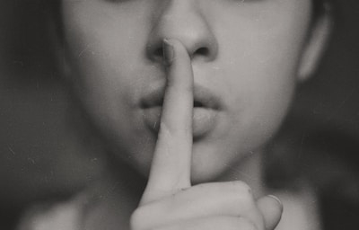 A person holds a finger to their lips in a gesture commonly associated with silence or secrecy. The image is in black and white, adding a classic or dramatic effect.