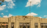 A weathered building facade with the text 'THE NEWS BROOKLYN GARAGE' displayed prominently. The building has a rustic and deteriorated look with visible grime and wear. Several large windows with rusted grilles are visible along the front, and a tall streetlight is positioned in the center of the image. The sky above is bright blue with scattered clouds.