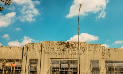 A weathered building facade with the text 'THE NEWS BROOKLYN GARAGE' displayed prominently. The building has a rustic and deteriorated look with visible grime and wear. Several large windows with rusted grilles are visible along the front, and a tall streetlight is positioned in the center of the image. The sky above is bright blue with scattered clouds.