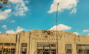 A weathered building facade with the text 'THE NEWS BROOKLYN GARAGE' displayed prominently. The building has a rustic and deteriorated look with visible grime and wear. Several large windows with rusted grilles are visible along the front, and a tall streetlight is positioned in the center of the image. The sky above is bright blue with scattered clouds.