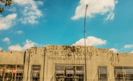 A weathered building facade with the text 'THE NEWS BROOKLYN GARAGE' displayed prominently. The building has a rustic and deteriorated look with visible grime and wear. Several large windows with rusted grilles are visible along the front, and a tall streetlight is positioned in the center of the image. The sky above is bright blue with scattered clouds.