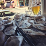 A tray filled with fresh oysters on ice is placed next to a glass of light beer, which sits on a window ledge. The background shows an outdoor urban scene with a red car and buildings visible through the window.