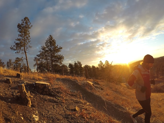 A person is jogging along a trail in a natural landscape during sunset. The trail is lined with dry grass and a few tree stumps. Tall pine trees are seen in the background against a sky filled with scattered clouds, and the sun is near the horizon, casting a warm glow over the scene.