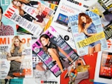 A variety of colorful magazines are scattered, featuring fashion models on the covers. Text in different languages and fonts highlights themes like fashion, beauty, and lifestyle.
