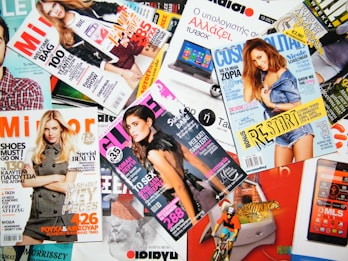 A variety of colorful magazines are scattered, featuring fashion models on the covers. Text in different languages and fonts highlights themes like fashion, beauty, and lifestyle.