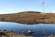 Wind turbines stand atop a gently sloping hill, overlooking a serene body of water that reflects their image. The landscape is characterized by a wide open sky and the earthy tones of the grassy terrain.