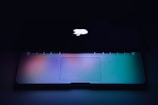 A partially open laptop with a glowing logo on the back of the screen, set against a dark background. The keyboard and touchpad are gently illuminated with a gradient of blue, green, and purple hues.