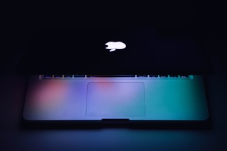 A partially open laptop with a glowing logo on the back of the screen, set against a dark background. The keyboard and touchpad are gently illuminated with a gradient of blue, green, and purple hues.