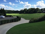 A serene landscape of a golf course featuring a well-maintained green field surrounded by trees under a vibrant blue sky dotted with fluffy white clouds. A white car is parked on a paved driveway bordered by neatly trimmed bushes. Golfers practice their swings in the distance.