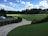 A serene landscape of a golf course featuring a well-maintained green field surrounded by trees under a vibrant blue sky dotted with fluffy white clouds. A white car is parked on a paved driveway bordered by neatly trimmed bushes. Golfers practice their swings in the distance.