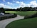 A serene landscape of a golf course featuring a well-maintained green field surrounded by trees under a vibrant blue sky dotted with fluffy white clouds. A white car is parked on a paved driveway bordered by neatly trimmed bushes. Golfers practice their swings in the distance.