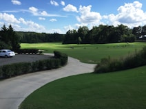 A serene landscape of a golf course featuring a well-maintained green field surrounded by trees under a vibrant blue sky dotted with fluffy white clouds. A white car is parked on a paved driveway bordered by neatly trimmed bushes. Golfers practice their swings in the distance.