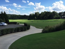 A serene landscape of a golf course featuring a well-maintained green field surrounded by trees under a vibrant blue sky dotted with fluffy white clouds. A white car is parked on a paved driveway bordered by neatly trimmed bushes. Golfers practice their swings in the distance.