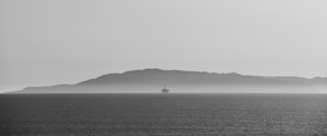 A stark black and white photograph capturing a solitary offshore oil rig against the backdrop of a distant, misty island across the water. The calm sea reflects the overcast sky, creating a serene yet somewhat isolated atmosphere.