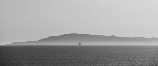 A stark black and white photograph capturing a solitary offshore oil rig against the backdrop of a distant, misty island across the water. The calm sea reflects the overcast sky, creating a serene yet somewhat isolated atmosphere.