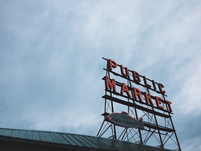 A large, vintage-style neon sign with the words 'Public Market' in bold red lettering, mounted on a metal frame. Below it, there's a smaller sign in the shape of a fish. The backdrop consists of a cloudy, overcast sky, contributing to a moody atmosphere, while the lower portion shows an angled green rooftop.