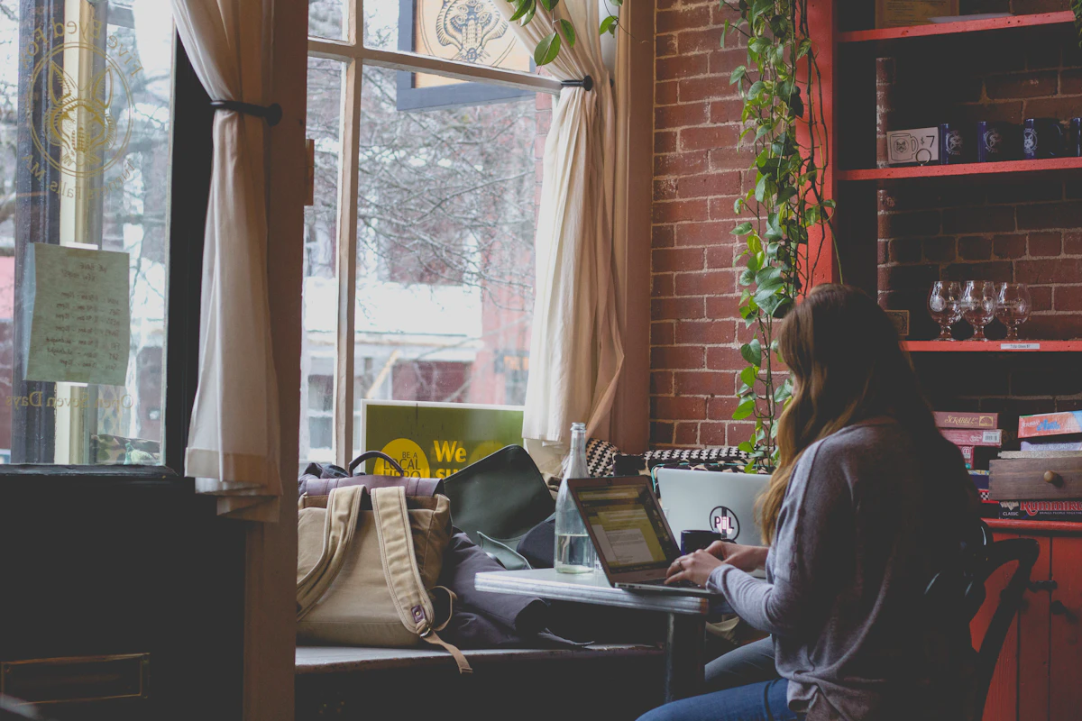 A woman works on a laptop at a window counter inside a shop with brick walls, shelves and a trailing plant