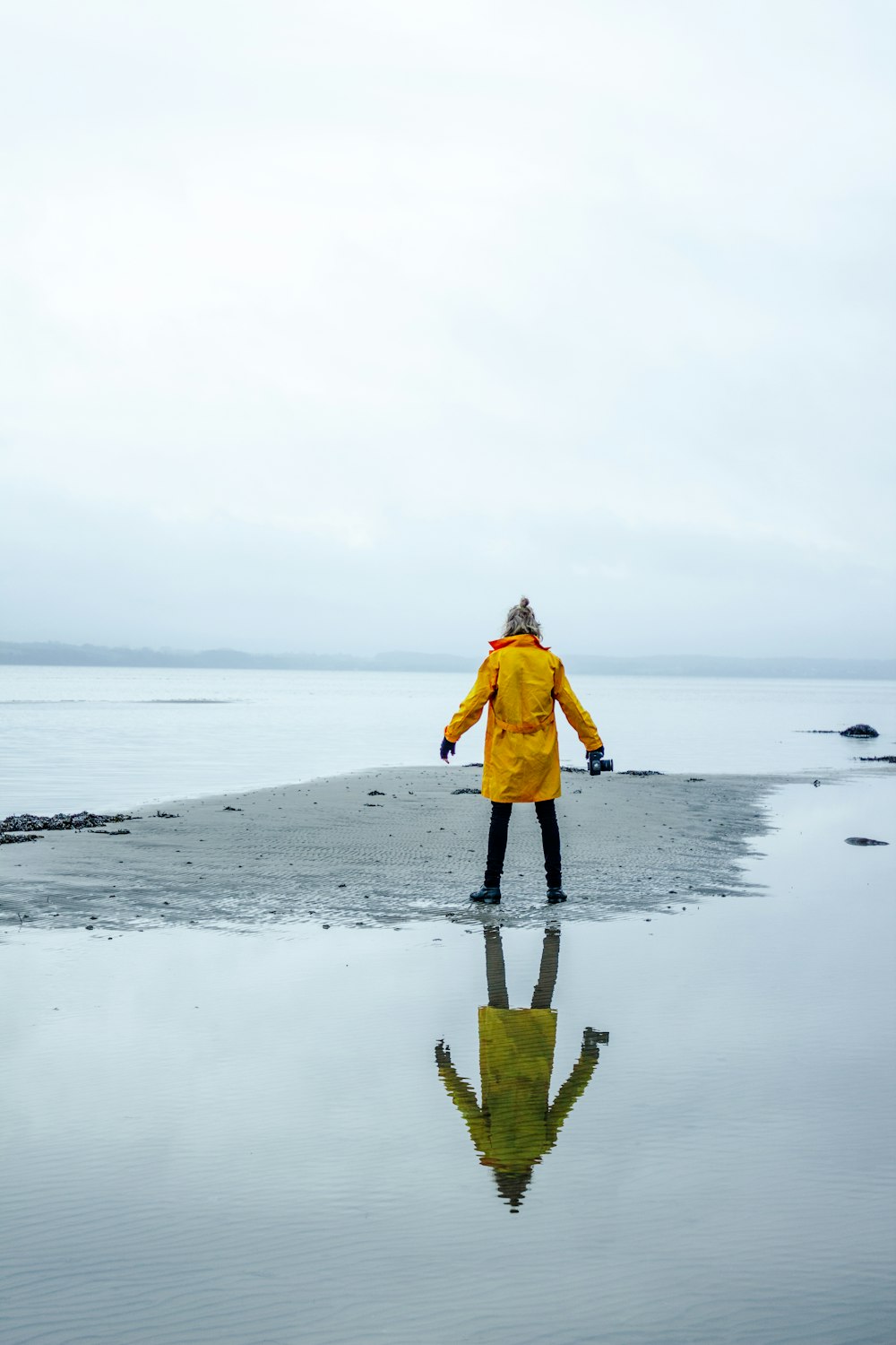 person in yellow coat on gray sand surrounded by water