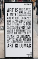 A black and white poster with various motivational phrases about art arranged in different fonts and sizes. Words include 'Art is love', 'Art is life', 'Art is a journey', among others. The poster has a minimalist design and is placed outside on what appears to be a city street.