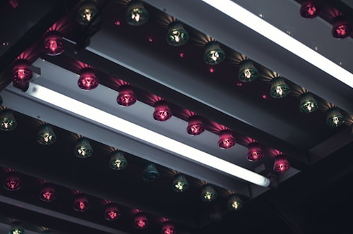 A series of light bulbs arranged in rows, featuring alternating pink and green colors, mounted on a reflective metallic surface with strips of LED fluorescent lighting. The setup creates an industrial and somewhat nostalgic ambiance.