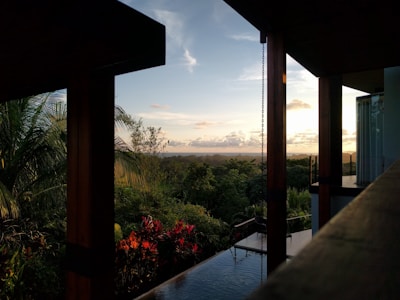 A tropical landscape view from the interior of a modern building with wooden beams. The scene includes lush green foliage, red flowers, and a clear sky with scattered clouds at sunset. A reflective water surface is partially visible, possibly part of an infinity pool.