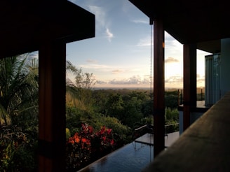 A tropical landscape view from the interior of a modern building with wooden beams. The scene includes lush green foliage, red flowers, and a clear sky with scattered clouds at sunset. A reflective water surface is partially visible, possibly part of an infinity pool.