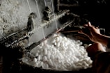 A pair of hands is working with a tool to unravel silk threads from white silkworm cocoons placed in steaming hot water. The industrial setup seems rustic, highlighting the traditional method of silk production.