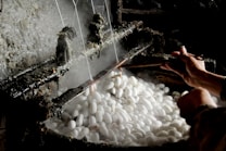 A pair of hands is working with a tool to unravel silk threads from white silkworm cocoons placed in steaming hot water. The industrial setup seems rustic, highlighting the traditional method of silk production.