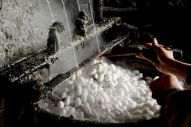 A pair of hands is working with a tool to unravel silk threads from white silkworm cocoons placed in steaming hot water. The industrial setup seems rustic, highlighting the traditional method of silk production.