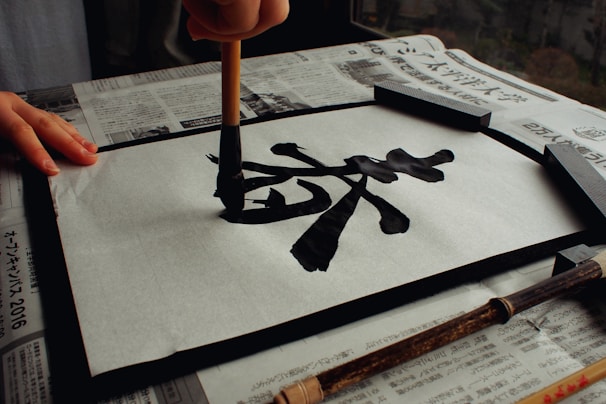 A hand holding a brush carefully paints black calligraphy characters on a piece of white paper. The paper is secured on a frame and rests on top of newspapers. A bamboo brush is positioned nearby.