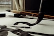 A close-up view of a traditional calligraphy brush making bold, black strokes on paper. The scene includes an additional brush and an ink block placed on the desk. Natural light filters through a window, casting soft shadows.