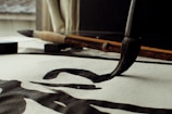 A close-up view of a traditional calligraphy brush making bold, black strokes on paper. The scene includes an additional brush and an ink block placed on the desk. Natural light filters through a window, casting soft shadows.
