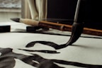 A close-up view of a traditional calligraphy brush making bold, black strokes on paper. The scene includes an additional brush and an ink block placed on the desk. Natural light filters through a window, casting soft shadows.