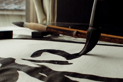 A close-up view of a traditional calligraphy brush making bold, black strokes on paper. The scene includes an additional brush and an ink block placed on the desk. Natural light filters through a window, casting soft shadows.