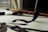 A close-up view of a traditional calligraphy brush making bold, black strokes on paper. The scene includes an additional brush and an ink block placed on the desk. Natural light filters through a window, casting soft shadows.