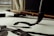 A close-up view of a traditional calligraphy brush making bold, black strokes on paper. The scene includes an additional brush and an ink block placed on the desk. Natural light filters through a window, casting soft shadows.