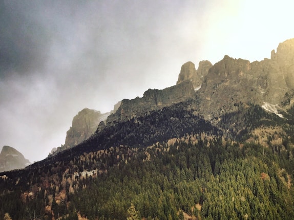 A dramatic mountain landscape with steep, jagged peaks rising above a dense forest of evergreen trees. The sky is filled with a mix of golden light and clouds, creating a moody and serene atmosphere.