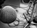 A close-up view of a silver microphone with a mesh grille, prominently featured on the left side of the image. The background includes tangled audio cables and a microphone stand, all in black and white.