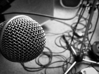 A close-up view of a silver microphone with a mesh grille, prominently featured on the left side of the image. The background includes tangled audio cables and a microphone stand, all in black and white.