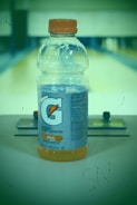 A plastic bottle of Gatorade Thirst Quencher, orange flavor, is positioned on a surface with a blurred background. The bottle has an orange cap, and detailed nutritional information is visible on the label.