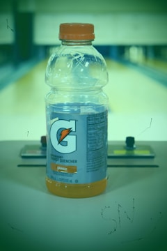 A plastic bottle of Gatorade Thirst Quencher, orange flavor, is positioned on a surface with a blurred background. The bottle has an orange cap, and detailed nutritional information is visible on the label.
