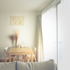 A minimalist dining space featuring light wood furniture, neutral-toned ceramics, and dried flowers in a glass vase.