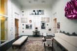 A well-decorated room featuring a variety of artwork on the walls, including photographs and framed art pieces. There is an elegant rug on the dark wooden floor, and the space is furnished with a white upholstered couch, a coffee table, and chairs. A vibrant magenta wall decoration adds a pop of color to the otherwise neutral color scheme. Soft lighting enhances the calm and sophisticated ambiance.